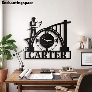 May include: A black wall clock featuring a silhouette of a construction worker holding a blueprint, integrated into a bridge-like structure. The clock face has gold hands and the name CARTER is displayed in bold capital letters at the bottom.