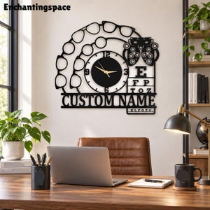 May include: A black metal wall clock featuring a circular clock face with gold hands, surrounded by silhouettes of various eyeglass frames and an optometry phoropter. The design includes an eye chart with the letters E, F, P, T, O, Z, ELFOTC and CUSTOM NAME.
