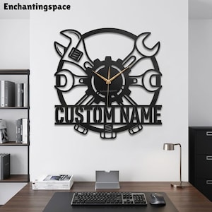 May include: A black wall clock featuring a circular gear design with crossed wrenches and a bolt at the bottom. The center of the clock has gold-colored hands and the text CUSTOM NAME is displayed in bold capital letters across the lower section.