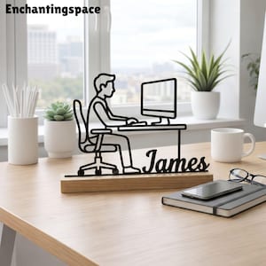 May include: A black metal silhouette of a person sitting at a desk with a computer monitor, mounted on a rectangular wooden base. The name James is written in black script lettering on the base. The object is placed on a wooden desk surface.