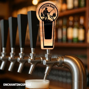 May include: A wooden beer tap handle with a circular design featuring a fisherman silhouette, a fish, and the words "CUSTOM NAME". The handle is part of a beer tap system with black handles. The text "ENCHANTINGSPACE" is visible.