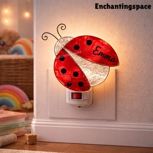 May include: A red and black ladybug night light plugged into a wall outlet. The ladybug has black spots and the name "Emma" is written on it. The night light has a decorative design and a switch.