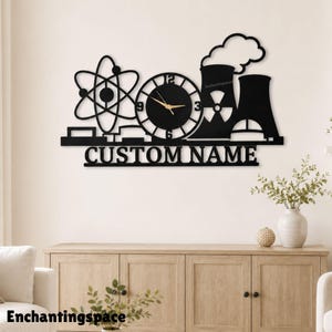 May include: A black metal wall clock featuring a central circular clock face with gold hands, flanked by an atomic symbol on the left and a nuclear power plant silhouette with smoke on the right. The text CUSTOM NAME is centered below.