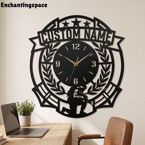 May include: A black circular wall clock featuring a soldier silhouette at the bottom, laurel leaf accents, and a banner at the top with the text CUSTOM NAME. The clock face has gold-coloured hands and white numerals at the 3, 6, 9, and 12 positions.