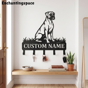 May include: A black metal wall-mounted key holder featuring a silhouette of a sitting dog positioned above a patch of grass. The design includes a rectangular plaque with the text CUSTOM NAME and five hooks for hanging keys, with some keys attached.