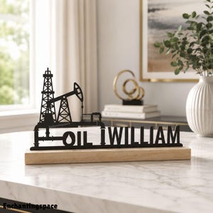 Op de afbeelding: Een zwart metalen uitsnede-bord met een silhouet van een boortoren en ja-knikker, geïntegreerd met de tekst OIL WILLIAM, gemonteerd op een rechthoekige lichtgekleurde houten voet. Het ontwerp bevat buisvormige structuren bij de apparatuur.
