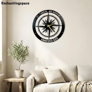 May include: A circular black wall clock featuring a central compass rose design. The outer ring displays the coordinates 27.7382 N, 82.6282 W and the text Coquina Key. The clock has thin gold-coloured hour and minute hands centred on the compass.