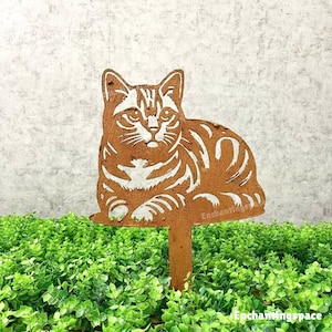 May include: A rusty brown metal cat garden stake. The cat is lying down with white stripes and details. The stake is in a bed of green plants. The text "Enchantingspace" is visible on the stake.
