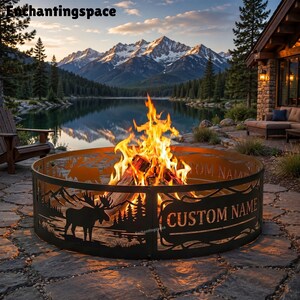 May include: A circular black metal fire pit ring featuring laser-cut silhouettes of a moose and pine trees. The metal ring includes a rectangular cutout area with the text CUSTOM NAME displayed in bold, capital letters, surrounding a bright fire.