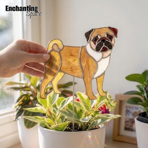 May include: A stained glass suncatcher shaped like a pug dog with tan, white, and black glass pieces held together by dark metal solder lines. The dog figure is attached to a thin metal stake, designed to be placed into a potted plant.