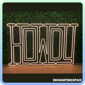 May include: A white neon sign with the word "HOWL" in a bold, blocky font. The sign is illuminated with a warm, yellow glow and is set against a backdrop of green foliage.