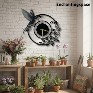 May include: A black circular wall clock featuring a hummingbird silhouette and floral accents. The clock face has thin white Roman numerals and gold-colored hands. The text Enchantingspace is visible in the upper right corner of the image.