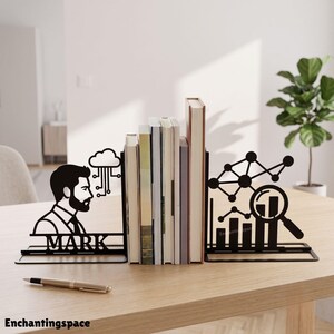 May include: A pair of black metal bookends featuring a silhouette of a person with a beard, a cloud icon, and the name MARK on one side, and a network diagram with a magnifying glass over a bar chart on the other, holding several books.