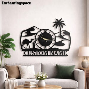 May include: A black wall clock featuring a desert landscape silhouette with a camel, a person, sand dunes, and a palm tree. The circular clock face has gold hands and numbers 3, 6, 9, and 12. Below the clock, the text CUSTOM NAME is displayed.