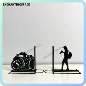 Puede incluir: Dos sujetalibros de metal negro con la silueta de una persona sosteniendo una cámara y un objetivo de cámara. Los sujetalibros están sobre una superficie blanca.