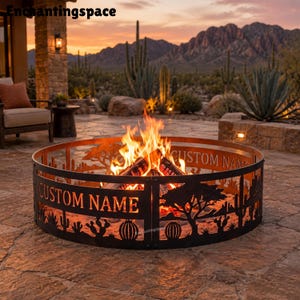 May include: A circular black metal fire pit ring featuring laser-cut silhouettes of desert cacti and acacia trees. The metal panels include the text CUSTOM NAME cut out of the design. Bright orange flames burn from wood logs inside the ring.