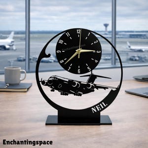 May include: Black airplane-themed desk clock with a circular clock face and gold hands. The clock features a cut-out design of an airplane and the name "NEIL". The clock is on a black base, with the text "Enchantingspace" at the bottom.