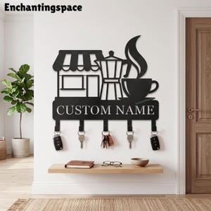 May include: A black metal wall-mounted key holder featuring a silhouette of a coffee shop storefront, a moka pot, and a steaming cup of coffee. Below the graphic, a rectangular plate displays the text CUSTOM NAME with five hooks for keys.