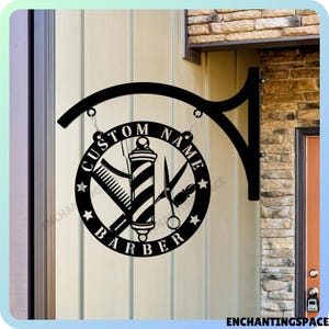 Puede incluir: Un letrero circular de barbero en metal negro con un gráfico central de poste de barbero flanqueado por un peine y unas tijeras. El texto CUSTOM NAME y BARBER está cortado en el marco circular, que cuelga de un soporte de pared negro.