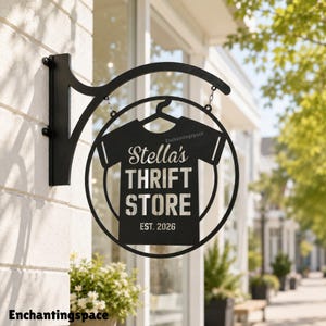 May include: A black metal wall-mounted sign featuring a circular frame and a central t-shirt silhouette. The text reads Stella's THRIFT STORE EST. 2026. The sign is suspended by chains from a curved bracket attached to a light-colored stone wall.