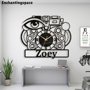 May include: Black wall clock featuring an eye and optical equipment design. The clock face is black with gold hands and the name "Zoey" below. The brand name "enchantingspace" is at the top.