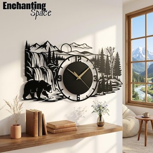 Pode incluir: Um relógio de parede de metal preto com um design de paisagem cortado a laser apresentando um urso, uma cascata, montanhas e pinheiros. O mostrador circular tem ponteiros dourados e os números 3, 6, 9 e 12. O texto Enchanting Space é visível.