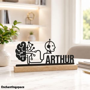 May include: A black metal desk sign featuring a stylized brain connected by circuit lines to a human head profile wearing headphones, with the name ARTHUR in bold capital letters, all mounted on a rectangular light wood base.