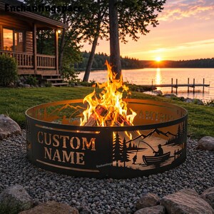 May include: A circular metal fire pit ring featuring laser-cut silhouettes of a person fishing in a boat, pine trees, and mountains. The side of the ring displays the text CUSTOM NAME in bold, capital letters, with a bright fire burning inside.