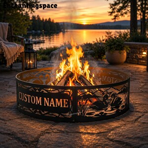 May include: A circular black metal fire pit ring featuring laser-cut silhouettes of swans on a lake with reeds and mountains. The front panel displays the text CUSTOM NAME in bold capital letters. A bright orange fire burns inside the ring.