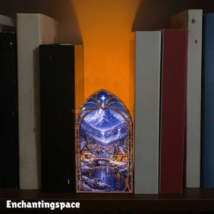 May include: A wooden, arched-shaped night light featuring a winter landscape with a mountain, river, and houses. The night light is illuminated, creating a warm glow. The text "Enchantingspace" is at the bottom.