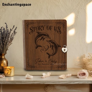 May include: Wooden book with the text "Story of Us" and two dolphins. The book is engraved with the names "John & Emily" and the date "Est. March 8, 2014". A heart-shaped lock secures the book.