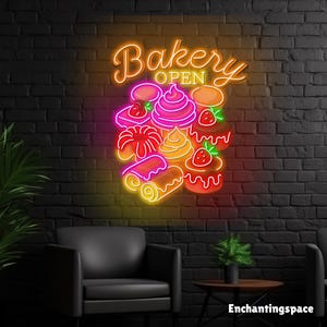 May include: A neon sign with the word "Bakery" in cursive and "Open" in block letters, with colorful illustrations of baked goods. The sign is mounted on a black brick wall, with a black chair and small table in front.