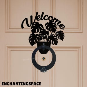 May include: Black metal door knocker featuring a potted plant design with large, stylized leaves and the word "Welcome" in a cursive font. The knocker includes a round handle and a small, decorative knob.