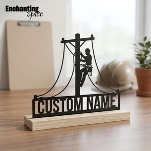 Puede incluir: Una silueta de metal negro de un trabajador eléctrico en un poste, montada sobre una base rectangular de madera clara. El diseño incluye líneas eléctricas y una sección recortada en la parte inferior con el texto CUSTOM NAME en mayúsculas.