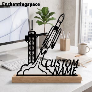 May include: A black laser-cut metal rocket ship launching from a launch tower, mounted on a rectangular light wood base. The design features a plume of smoke at the base and the text CUSTOM NAME in a bold, sans-serif font on the right side.