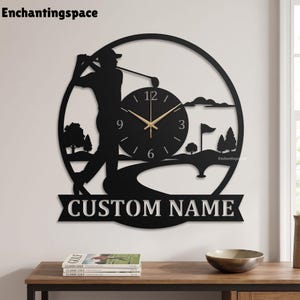 May include: A circular black wall clock featuring a silhouette of a golfer swinging a club on a course. The clock face has gold hands and numbers 3, 6, 9, and 12. Below the circular frame is a banner with the text CUSTOM NAME in white letters.