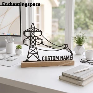 Könnte beinhalten: Eine schwarze Metallsilhouette eines Strommasts mit seitlich verlaufenden Stromleitungen, montiert auf einem rechteckigen Holzsockel. Der Text CUSTOM NAME ist in einer fetten, serifenlosen Schrift am unteren Teil des Strommasts ausgeschnitten.