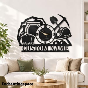 May include: A black metal wall clock featuring a mining theme with a mine entrance, a cart, a shovel, a pickaxe, and a hard hat. The clock face has gold hands and the text CUSTOM NAME integrated into the design below the central clock mechanism.