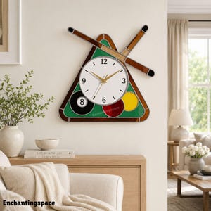 May include: A wall clock shaped like a billiard rack with two crossed cues. The clock face is white with black numbers and gold hands. It features a black eight ball, a red ball, and a yellow ball set against a green background with brown borders.