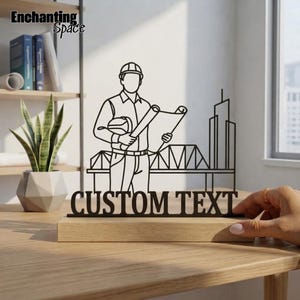 Pode incluir: Uma silhueta de metal preto de um engenheiro com capacete e plantas, em frente ao contorno de uma ponte e um edifício. A peça apresenta as palavras CUSTOM TEXT em letras negrito e está montada numa base de madeira retangular.