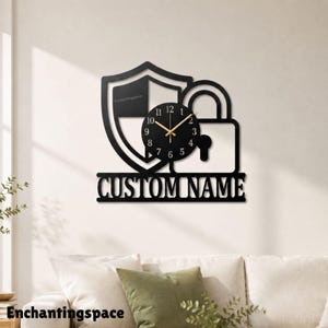 Könnte beinhalten: Eine schwarze Wanduhr mit einem Schild- und Vorhängeschloss-Design. Das runde Zifferblatt hat weiße Zahlen und goldene Zeiger. Unter der Uhr befindet sich der Schriftzug CUSTOM NAME in fetten, schwarzen Buchstaben auf einem horizontalen Balken.