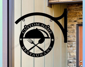 Pode incluir: Uma placa suspensa circular de metal preto com a silhueta de uma campânula de comida, um garfo e uma colher. O texto CUSTOM NAME e RESTAURANT está recortado ao longo da borda, com duas pequenas estrelas separando as palavras no círculo.