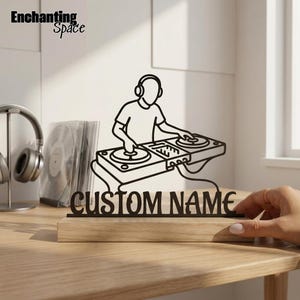Puede incluir: Una silueta de metal negro de un DJ con auriculares operando una mesa de mezclas, montada sobre una base rectangular de madera clara. El texto CUSTOM NAME está recortado en letras de metal negro en la parte inferior del diseño.