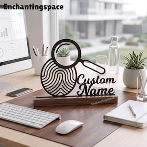 May include: A black laser-cut sign featuring a magnifying glass icon over a fingerprint pattern and the text Custom Name, mounted on a rectangular dark wood base. The sign is positioned on a desk with a computer, keyboard, and mouse.