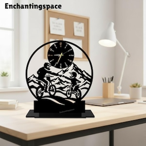 Könnte beinhalten: Eine schwarze, runde Tischuhr mit der Silhouette von zwei Personen auf Dirtbikes, die vor Berggipfeln die Fäuste aneinanderstoßen. Das Zifferblatt hat weiße Zahlen und goldene Zeiger. Der Sockel ist ein schwarzer, rechteckiger Ständer mit Text.