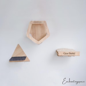 May include: Three wooden cat shelves mounted on a white wall. The shelves are in different shapes, including a triangle, a hexagon, and a rectangle. The shelves are made of natural wood and have a soft, gray fabric lining. The rectangular shelf has the word "Garfield" engraved on it.