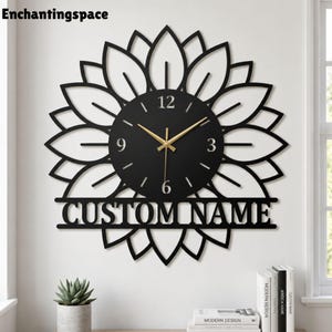 May include: A black wall clock shaped like a sunflower with gold-colored hands. The central circular face features the numbers 3, 6, 9, and 12. Below the center, a horizontal bar displays the text CUSTOM NAME in capital letters.