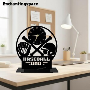 Op de afbeelding: Een zwarte ronde bureauklok met een honkbalthema, voorzien van een handschoen, twee gekruiste knuppels en een helm. De wijzerplaat heeft gouden wijzers en cijfers. Onder de klok staat de tekst BASEBALL DAD in een vetgedrukt lettertype.