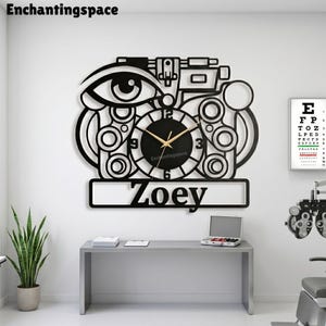 May include: A black wall clock shaped like an optometrist's phoropter, featuring a central circular clock face with gold hands, a stylized eye graphic, and a rectangular nameplate at the bottom with the name Zoey printed in black lettering.