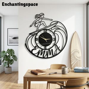 May include: A black wall clock featuring a cutout design of a person surfing on a wave. The clock face is circular with a black background, white numerals at 3, 6, 9, and 12, and gold hands. The name Emma is integrated into the lower frame.